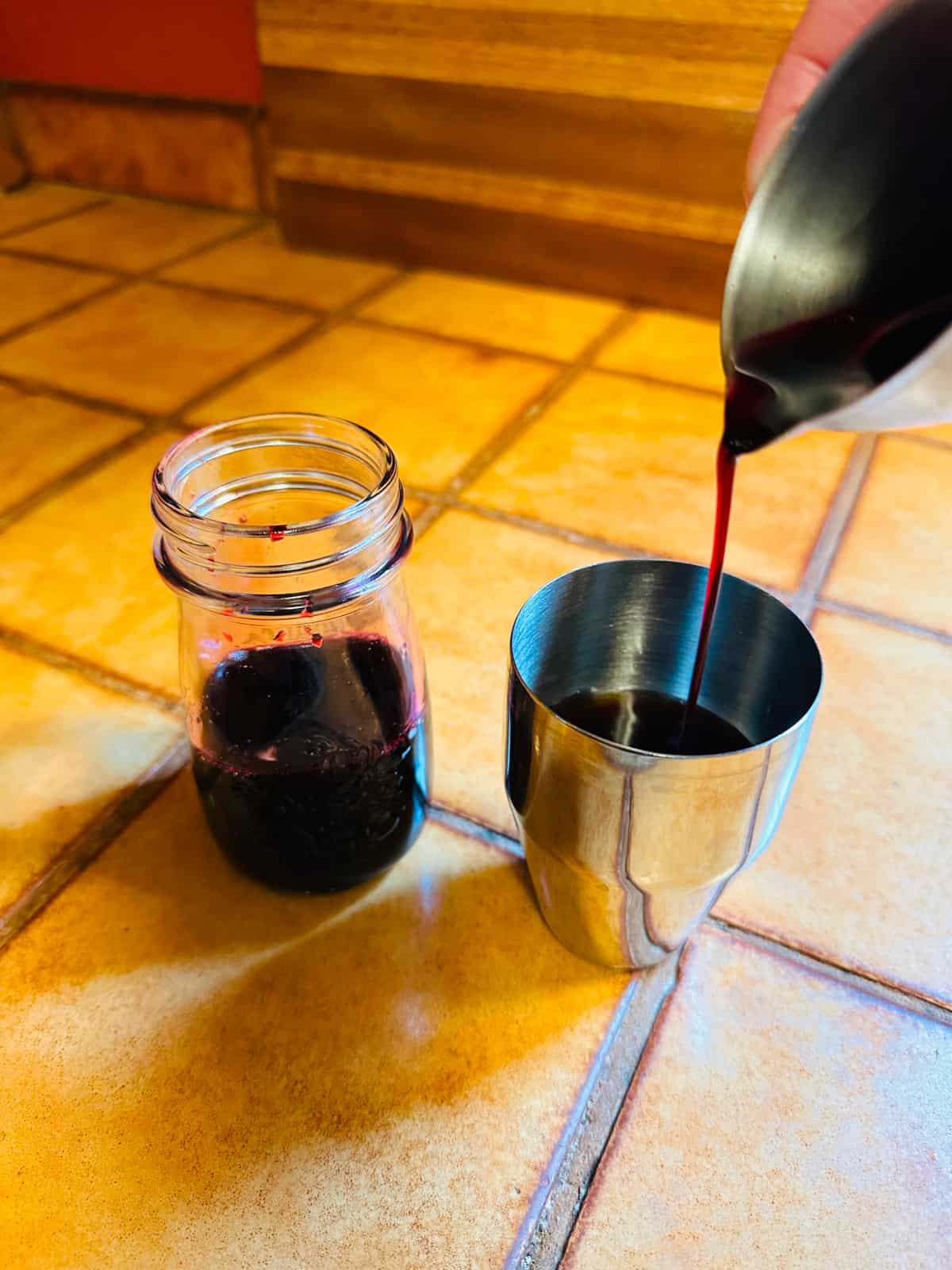 Dark violet liquid being poured from a steel measuring jigger into a small steel cocktail shaker next to a small bottle of dark violet liquid.