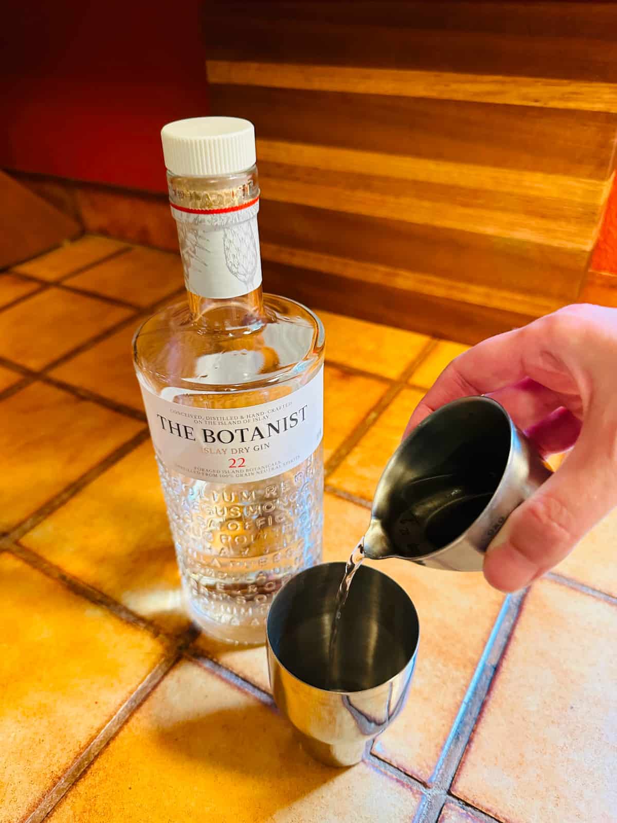 Clear liquid being poured from a steel measuring jigger into a small steel cocktail shaker next to a bottle of Botanist gin.