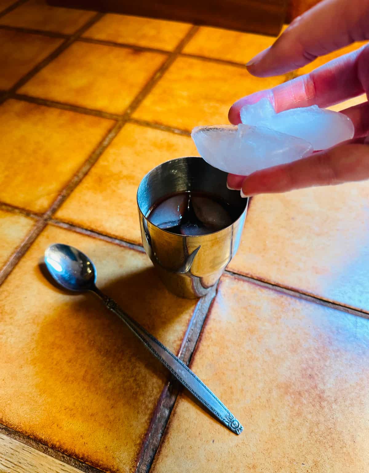 Ice cubes being dropped into a small steel cocktail shaker sitting on a brown tile counter next to a long metal spoon.