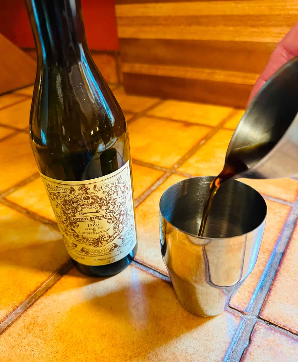 Brown liquid being poured from a steel measuring jigger into a small steel cocktail shaker next to a bottle of Carpano Antica sweet vermouth.