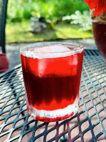 Dark red liquid and large ice cube in an old fashioned glass sitting on a lattice style black metal table with greenery in the background.