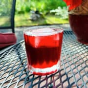 Dark red liquid and large ice cube in an old fashioned glass sitting on a lattice style black metal table with greenery in the background.