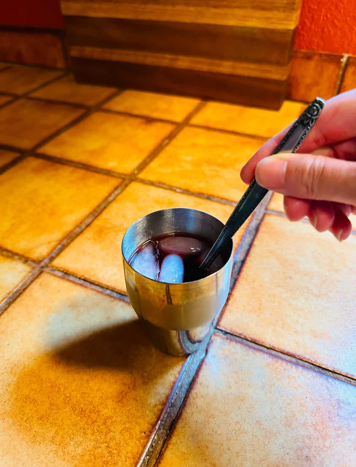 Dark red liquid with ice being stirred with a long metal spoon in a small steel cocktail shaker sitting on a brown tile counter.