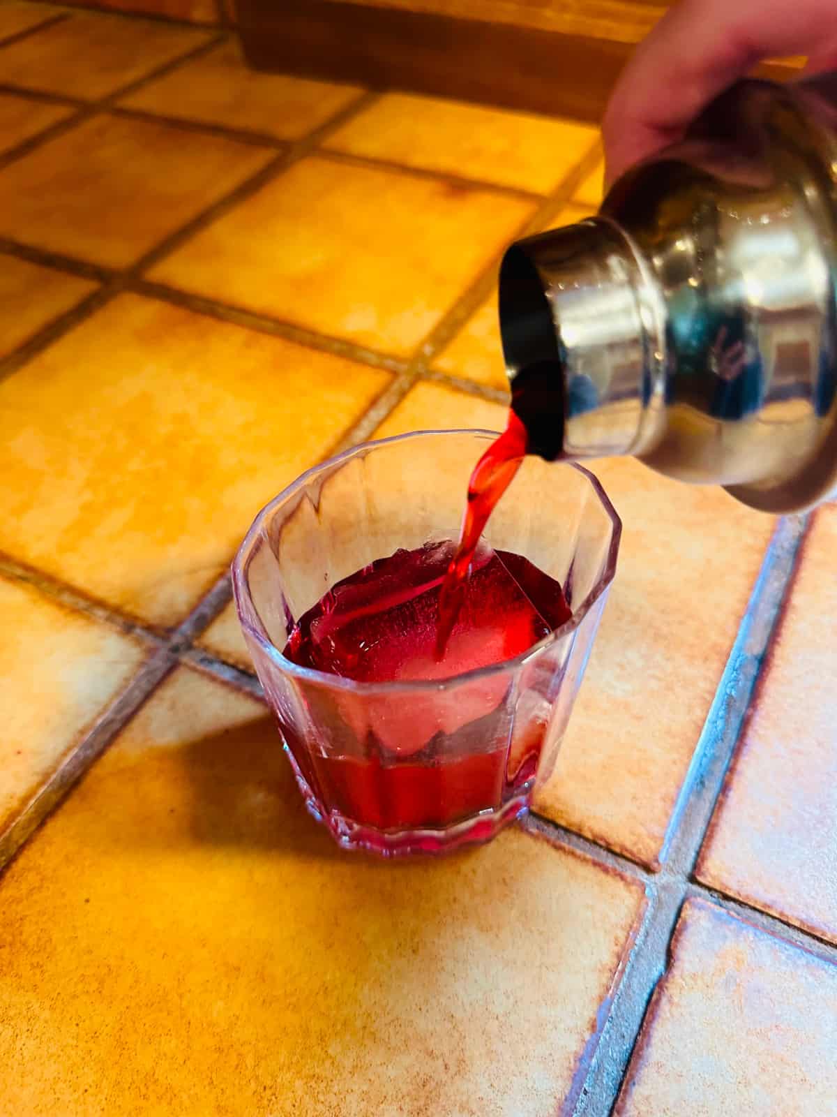 Red liquid being poured from a steel cocktail shaker over a large ice cube in an old fashioned glass sitting on a brown tile counter.