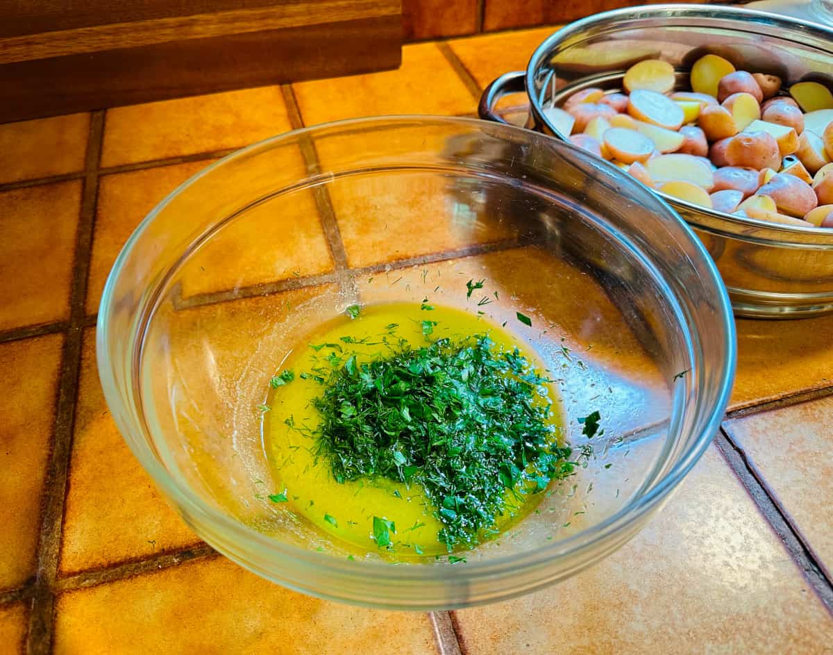 Chopped dill and parsley sitting on top of a shallow pool of yellow liquid in a glass bowl next to a steamer basket full of potatoes.