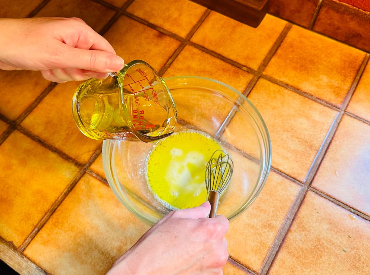 Olive oil being poured from a glass measuring cup into a shallow pool of yellow liquid being whisked in a glass bowl.