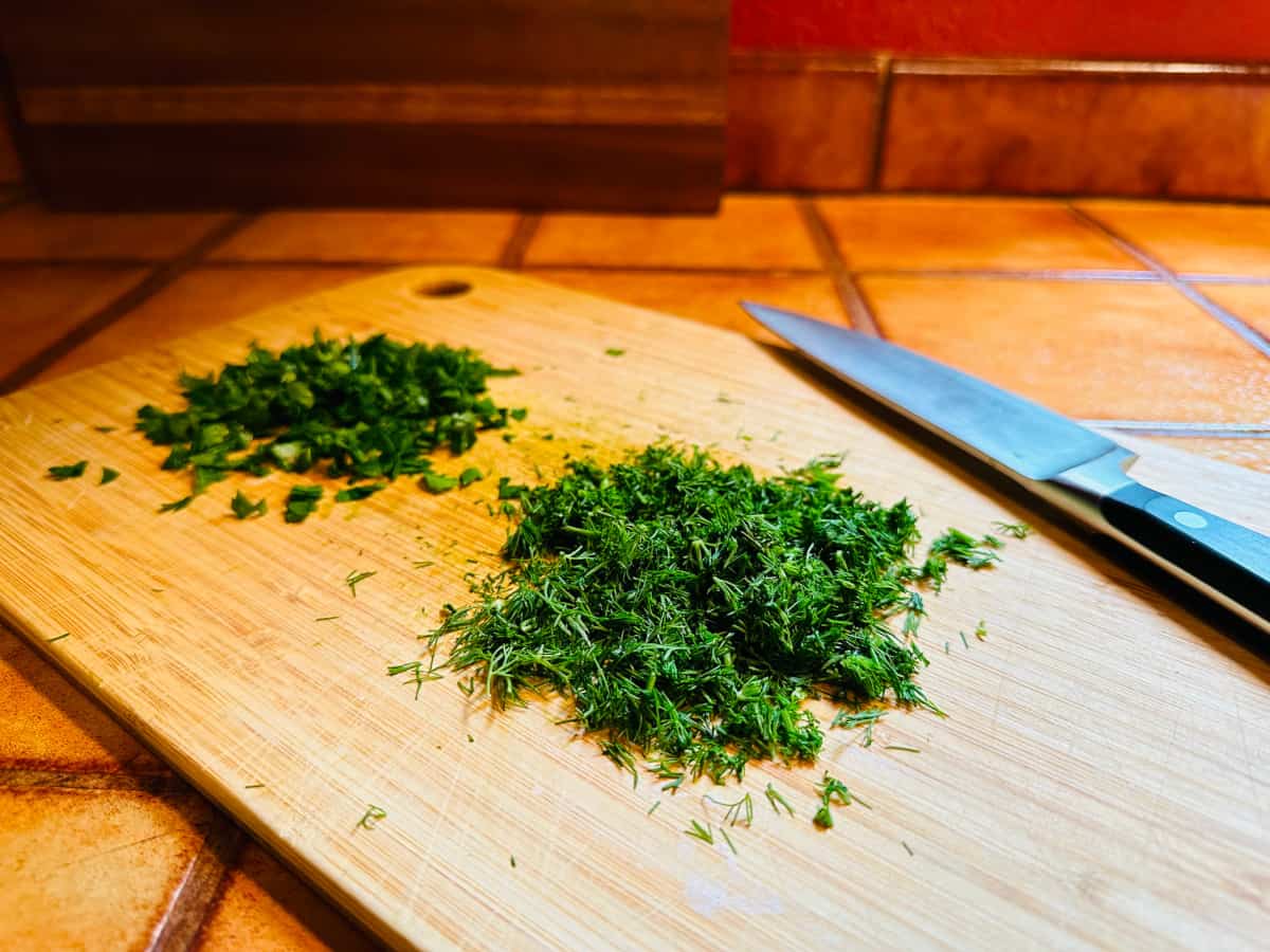 A heap of chopped dill and a heap of chopped parsley on a cutting board with a chef's knife.