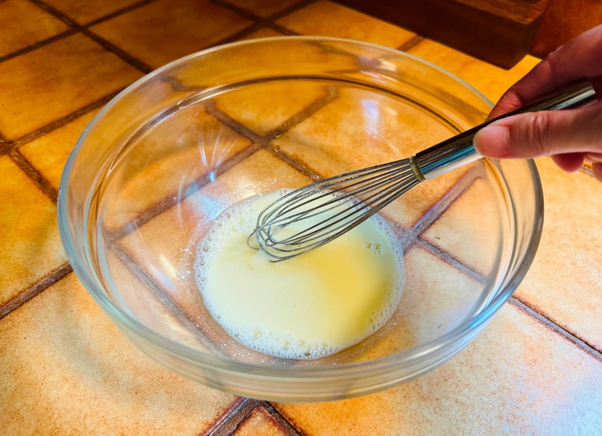 Opaque yellow liquid being mixed with a small steel whisk in a glass bowl.