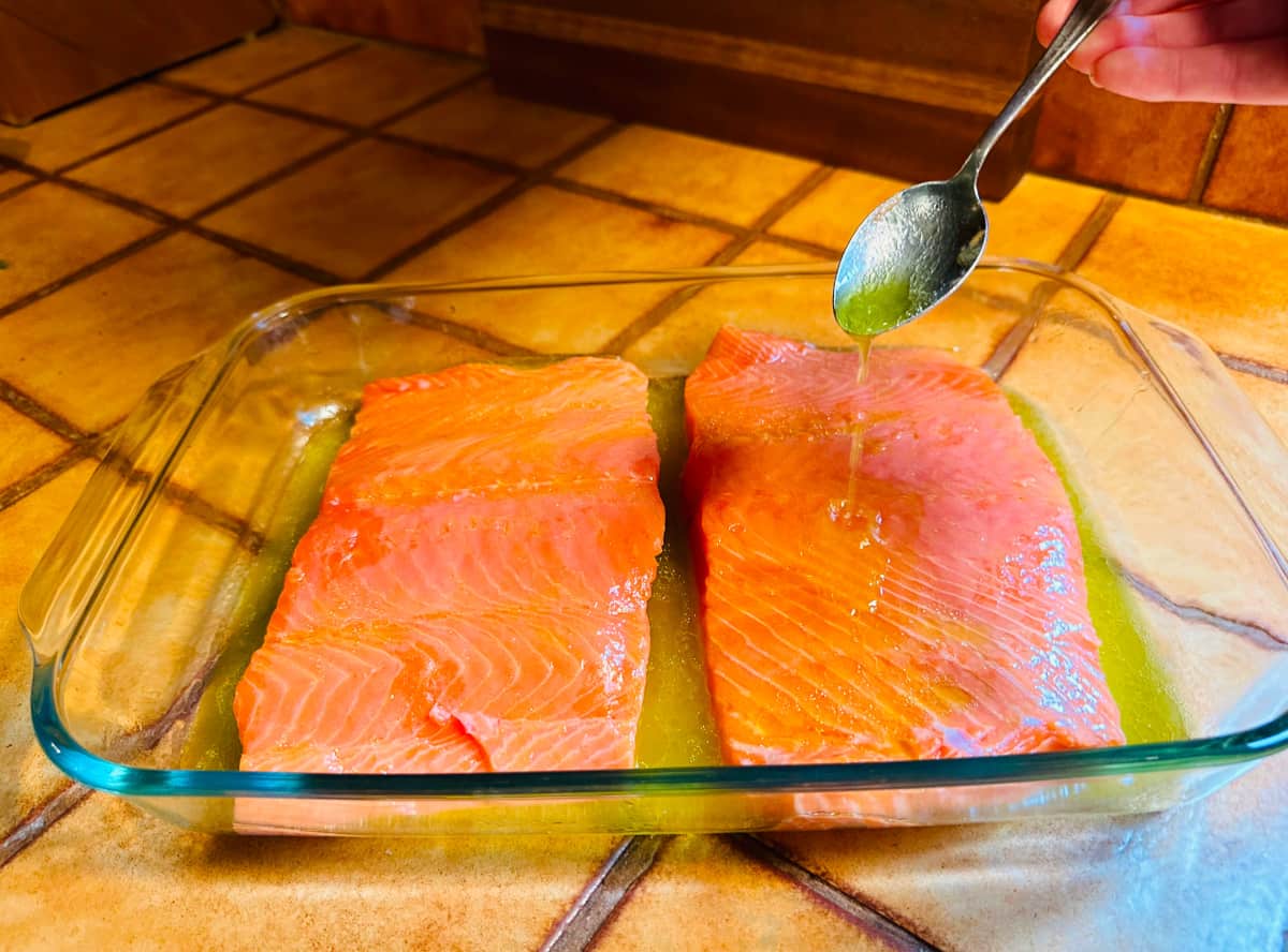 Raw salmon fillet in a large glass roasting dish being basted in lemon juice and olive oil with a spoon.