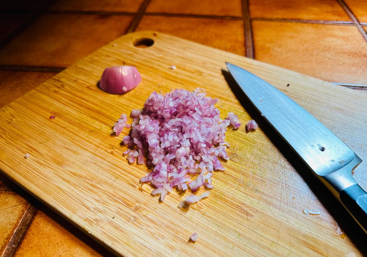 Minced shallot on a cutting board between a chunk of shallot and a chef's knife.
