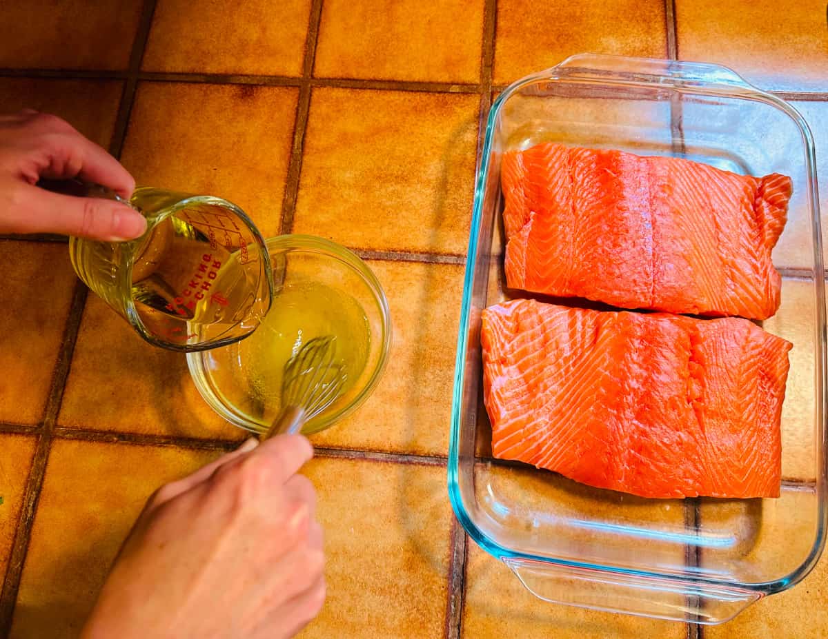 Olive oil being poured from a glass measuring cup and whisked into a small bowl of lemon juice next to a glass roasting dish with raw salmon fillet.