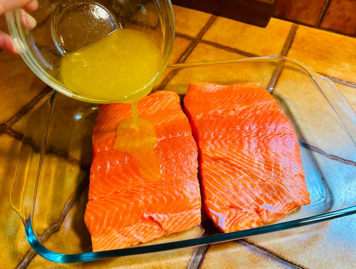 Lemon juice and olive oil being poured from a small glass bowl over raw salmon fillet in a large glass roasting dish.