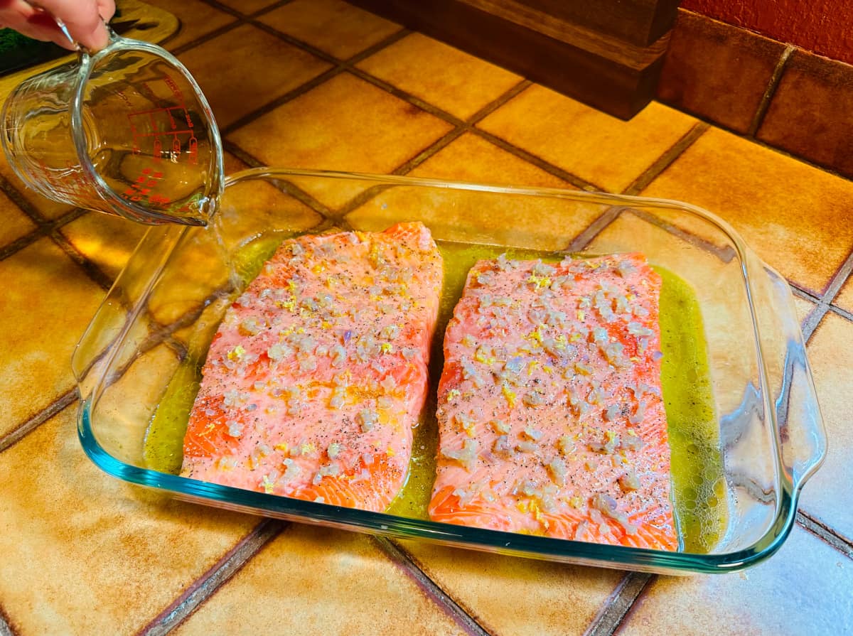 White wine being poured from a glass measuring cup around raw salmon fillet sprinkled with sautéed shallot and lemon zest in a large glass roasting dish with a shallow pool of lemon juice, olive oil, and wine in the bottom.