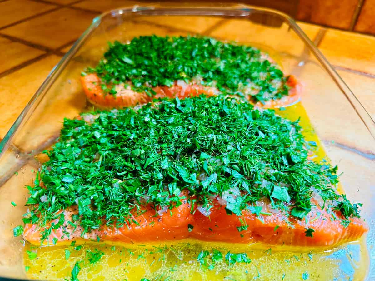 Raw salmon fillet covered with roughly chopped dill and parsley in a glass roasting dish with a shallow pool of lemon juice, olive oil, and wine in the bottom.