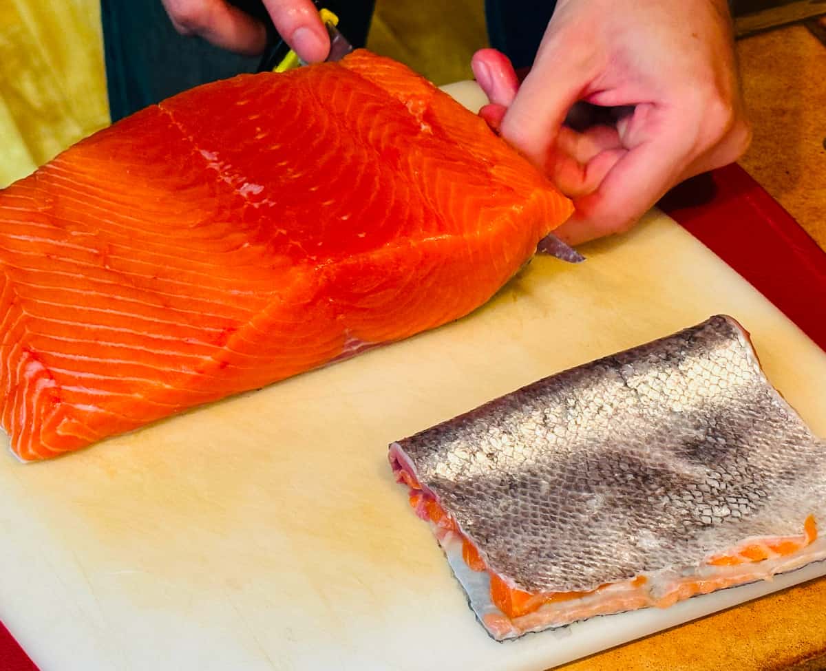 Raw salmon fillet being skinned with a knife next to a silver piece of salmon skin.
