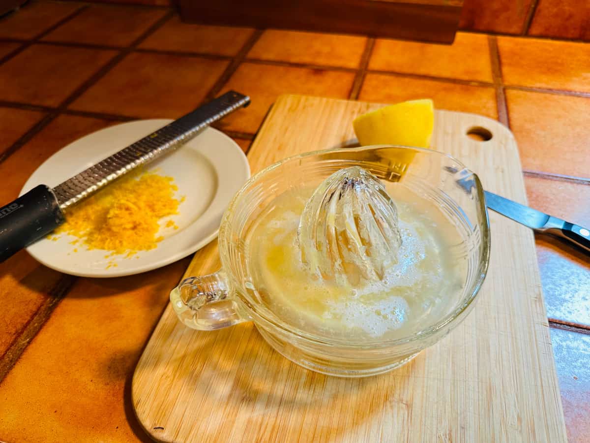 Glass juicer containing lemon juice siting on a cutting board with a paring knife and a lemon half next to a small white plate with lemon zest and a microplane grater.
