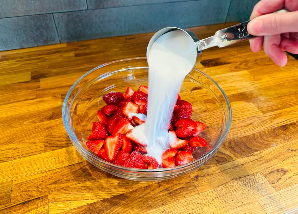 Sugar being poured from a metal measuring cup over sliced strawberries in a glass bowl.