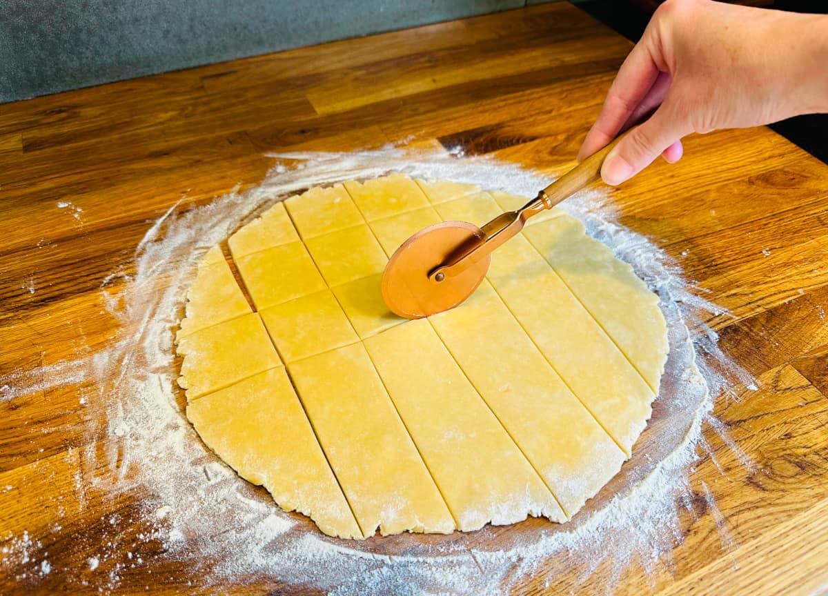 Rolled out circle of pastry on a floured wooden surface being cut into rough squares with a copper pizza cutter.