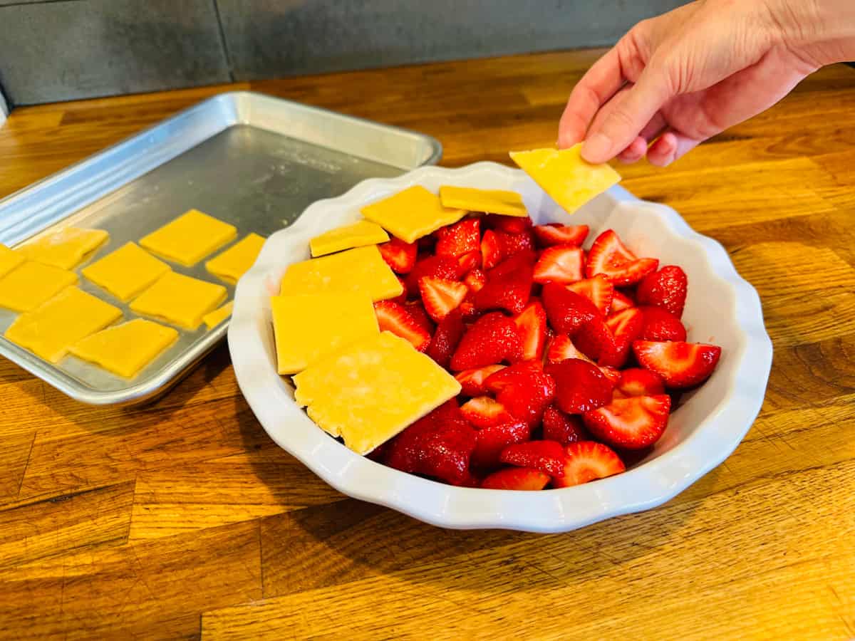 Squares of pastry dough being placed on top of sliced strawberries in a white ceramic pie plate.