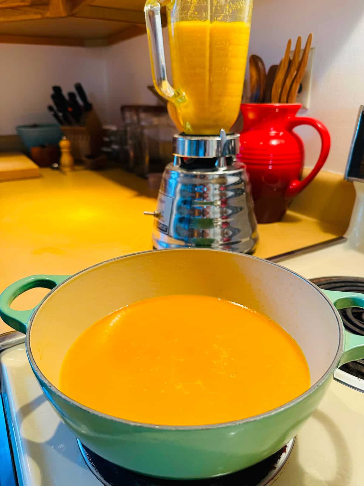 Puréed creamy carrot soup in a green pot with a blender behind it. 