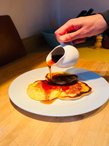 Hand pouring syrup from white pitcher over pancakes on white plate.