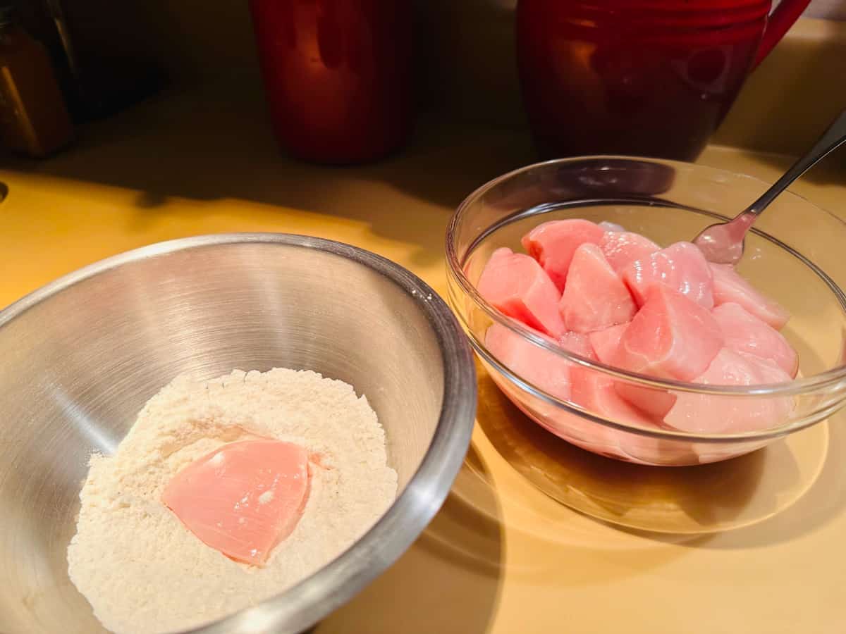 Bowl of flour next to bowl of raw chicken chunks.