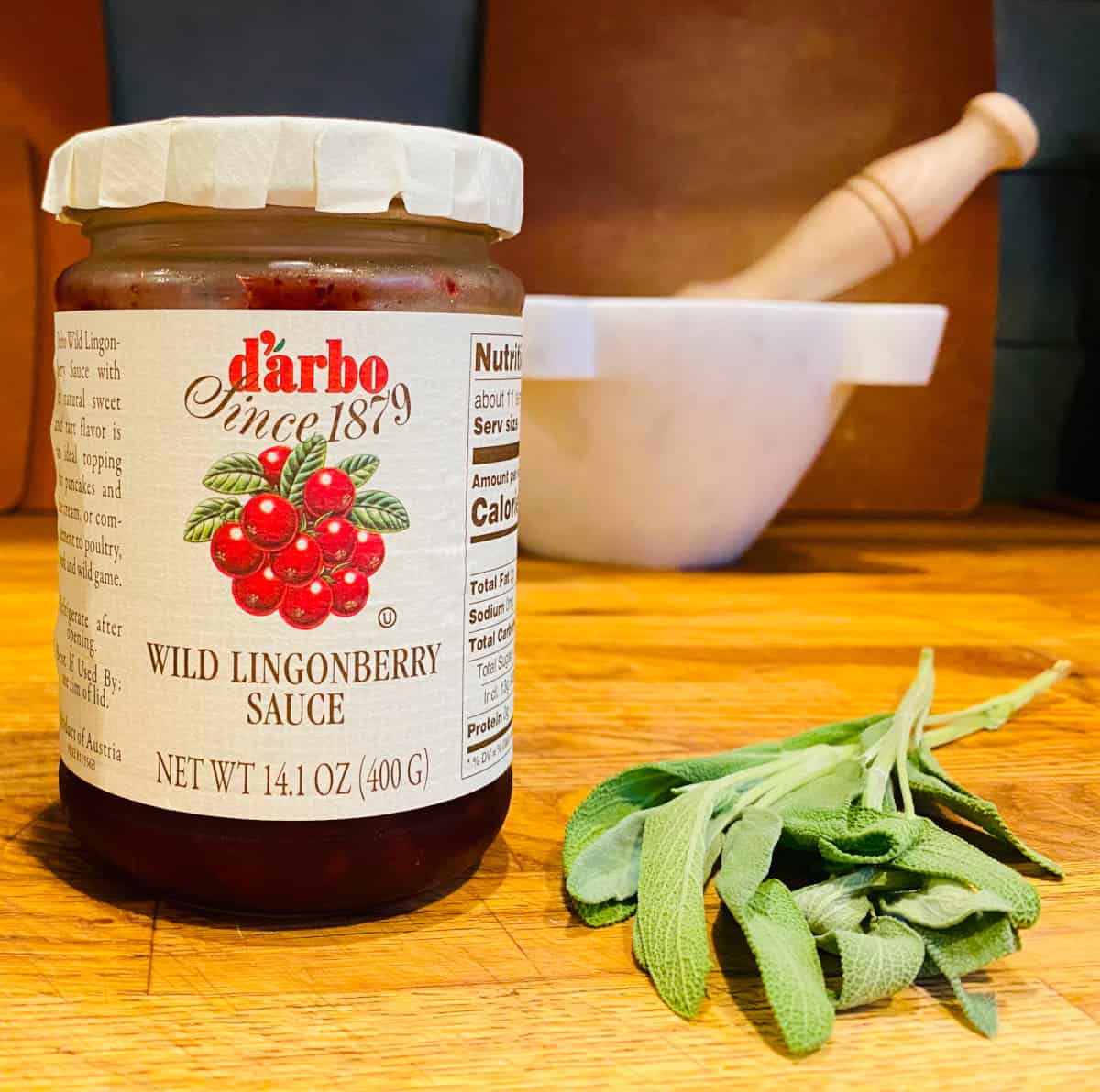 Jar of wild lingonberry sauce and small bunch of fresh sage on a wooden counter.