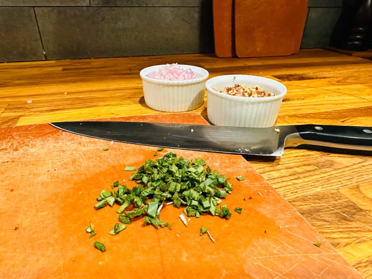 Chopped sage on a cutting board with a butcher knife and two white ramekins filled with chopped shallots and chopped pecans.