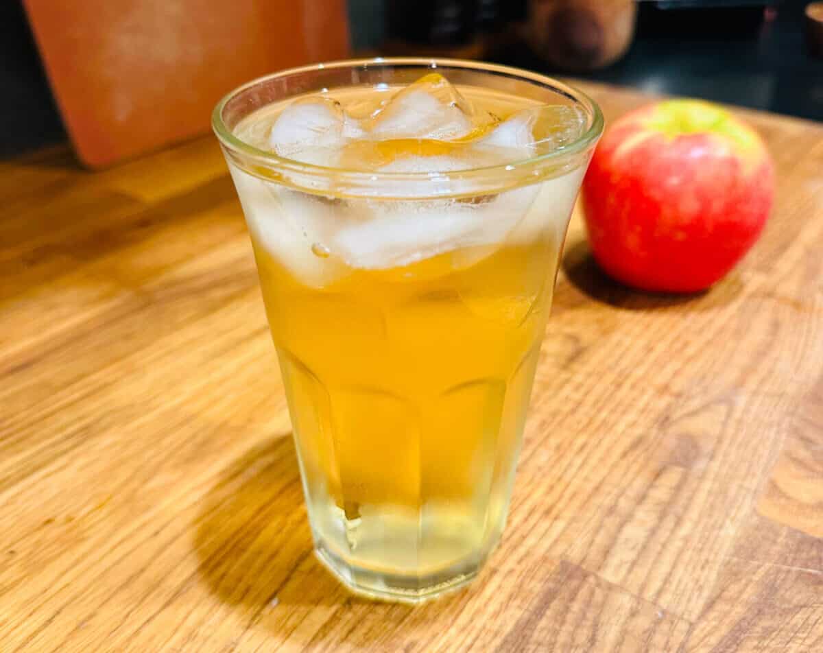Yellow liquid and ice in a clear glass next to an apple.