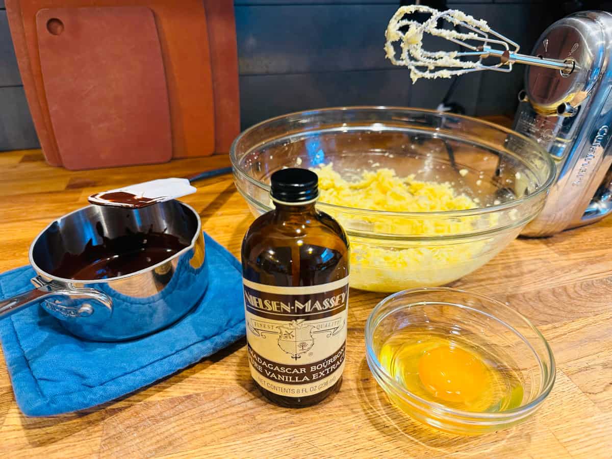 Saucepan of melted chocolate, bottle of vanilla extract, cracked egg in small glass bowl, and cookie batter in large glass bowl