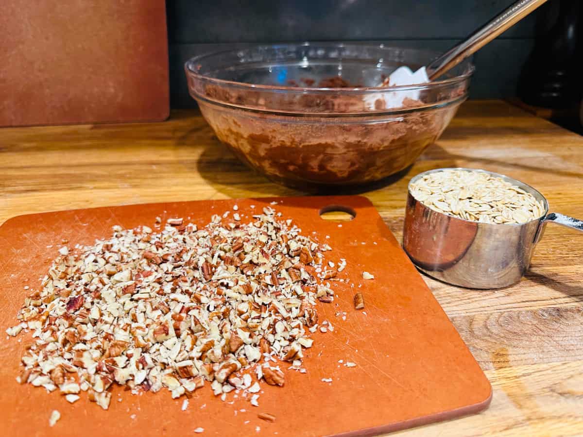 Chopped pecans on a cutting board, measuring cup of oats, and glass bowl of chocolate cookie batter