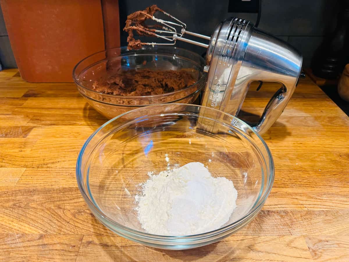 Glass bowl of dry ingredients next to bowl of chocolate cookie batter with electric mixer