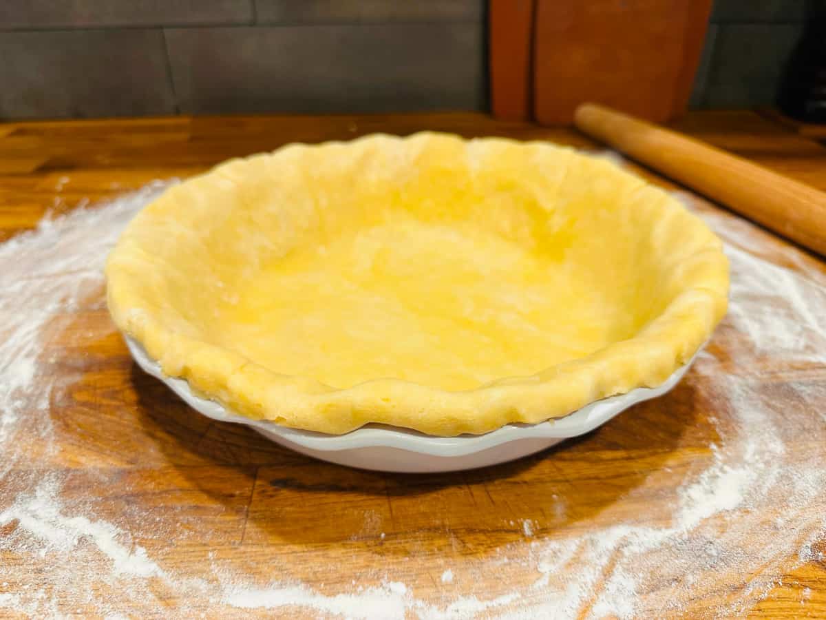 Butter pie dough in a white pie dish next to a rolling pin.