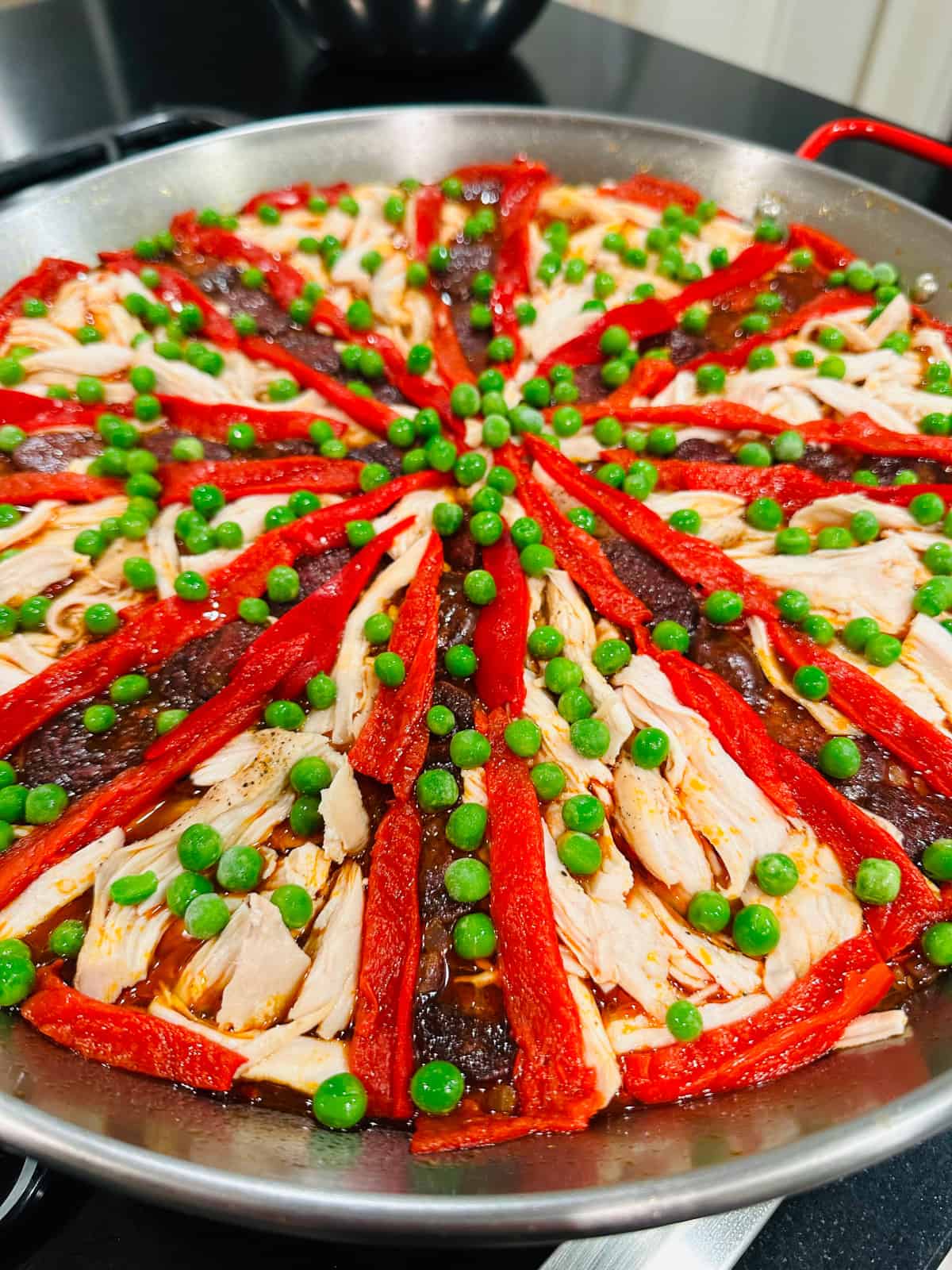 Chicken, chorizo, piquillo peppers, and peas arranged like spokes of a wheel in metal pan.