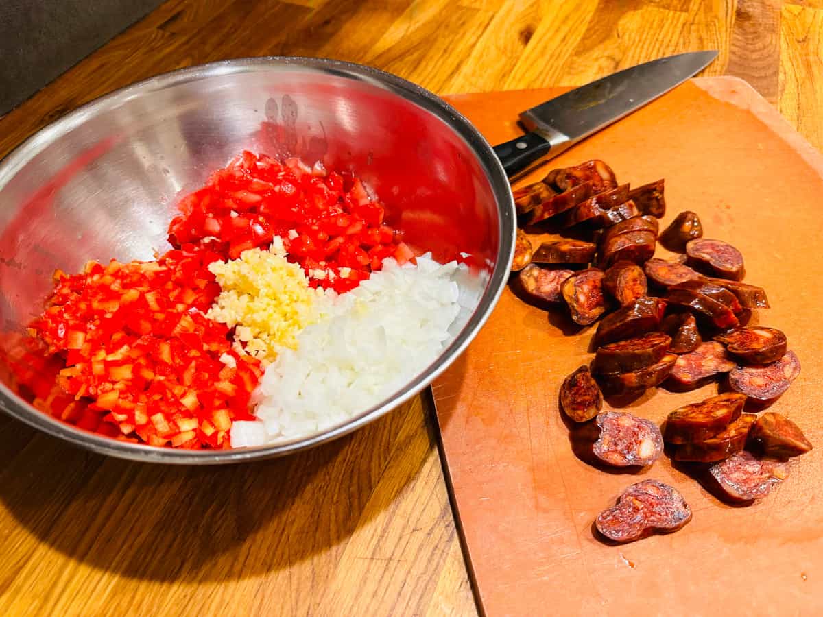 Steel bowl containing chopped vegetables for sofrito next to chopped chorizo on a cutting board.