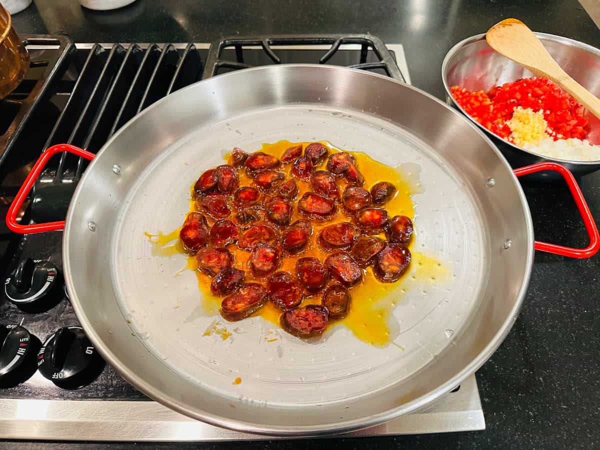 Sliced chorizo frying in a metal pan with red handles.