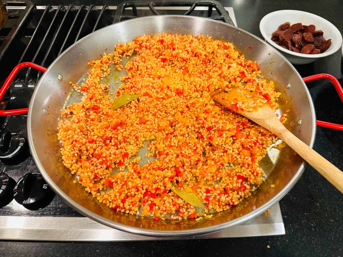Rice, spices, and sofrito mixed together in a metal pan with red handles and a wooden spatula.