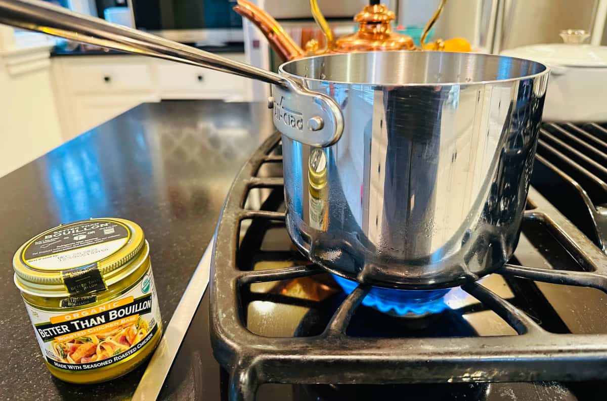 Jar of chicken bouillon next to small steel saucepan heating on stove.