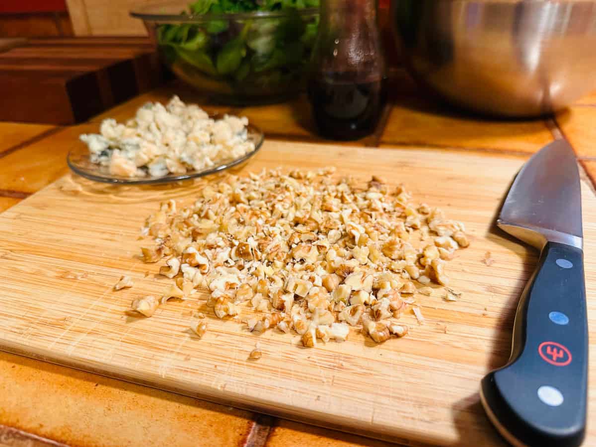 Chopped walnuts on a wooden cutting board with plate of blue cheese crumbles.