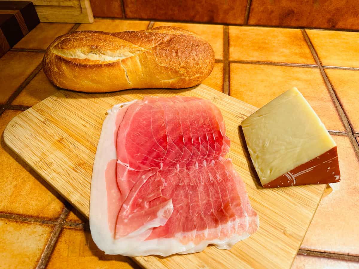 Loaf of bread, several slices of prosciutto, and wedge of fontina on wood cutting board.