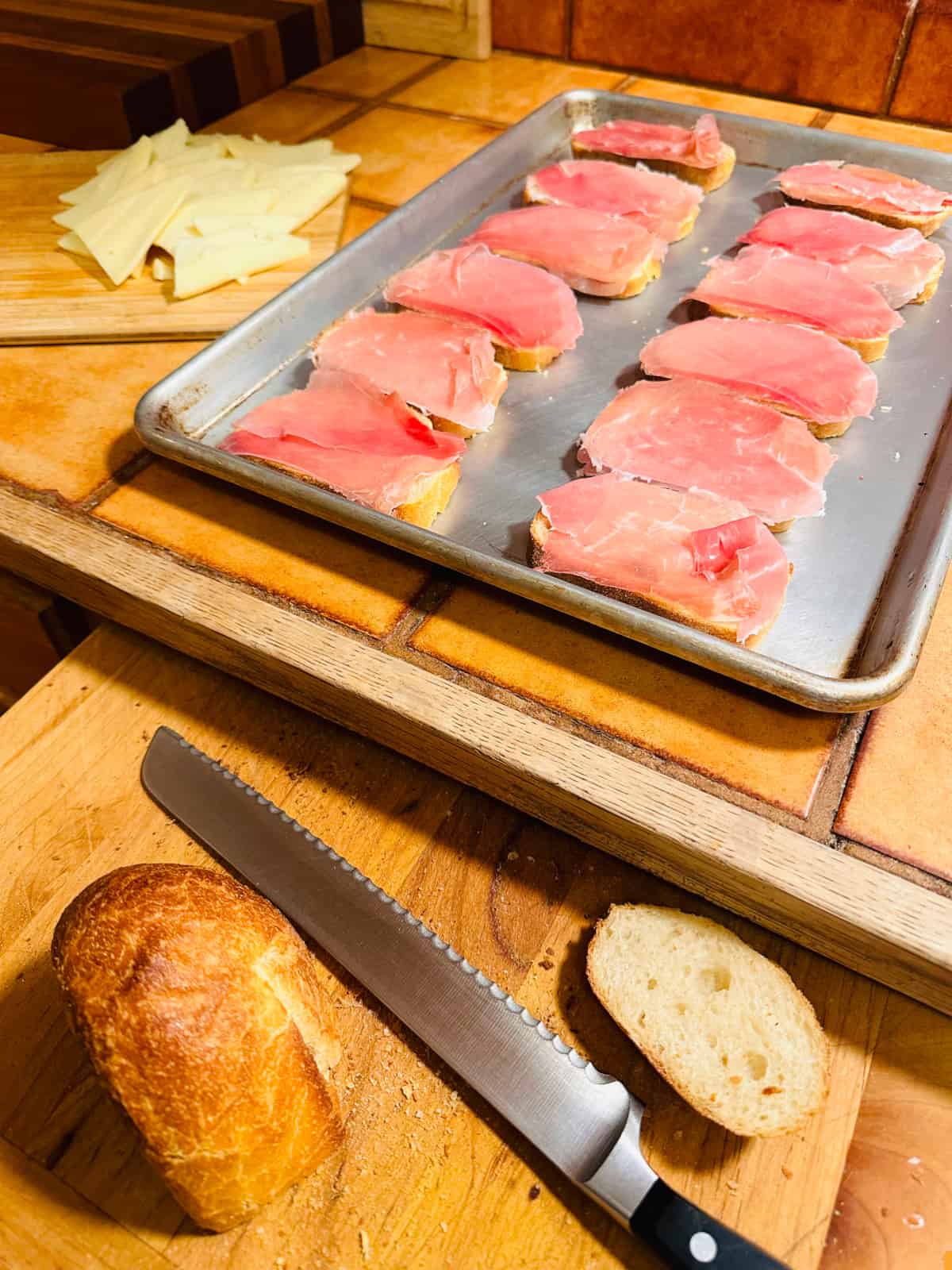 Bread heels and bread knife on cutting board in front of metal baking sheet holding slices of bread covered with prosciutto.