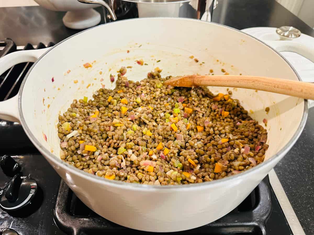 Lentils mixed with cooked vegetables in a white pot on the stove with a wooden spoon.