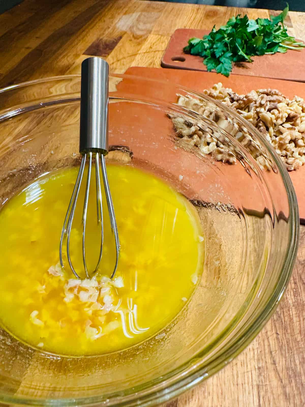 Yellow colored dressing in a glass bowl with a metal whisk in front of walnuts and parsley on wood cutting boards.