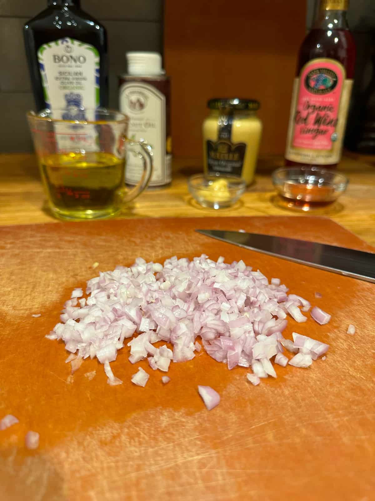 Chopped shallots on a wood cutting board in front of dressing ingredients for French lentil salad with feta.