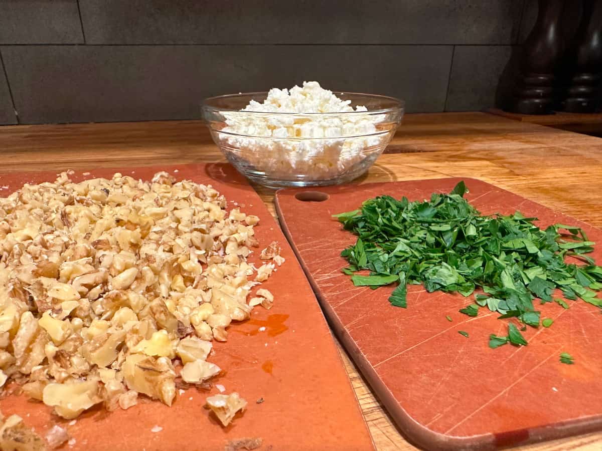 Chopped walnuts and chopped parsley on wood cutting boards in front of a glass bowl of crumbled feta.