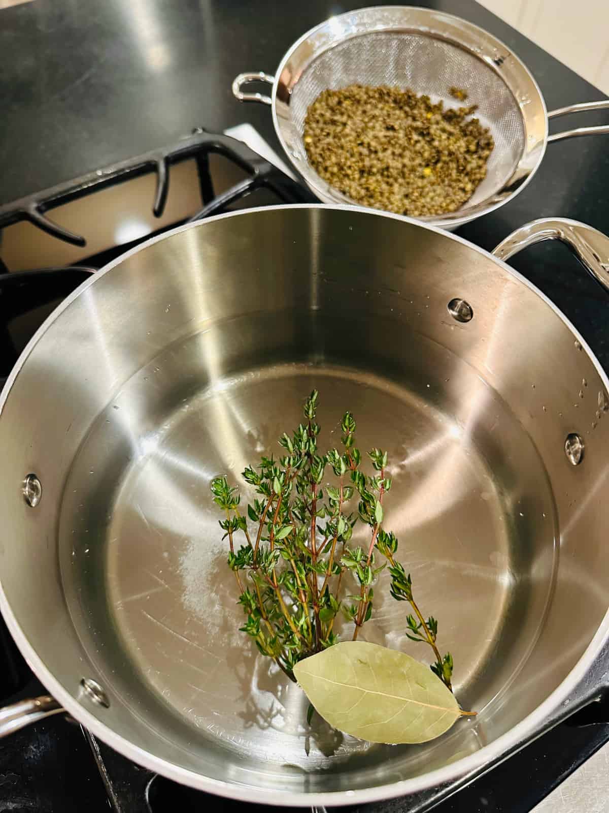 Thyme sprigs and bay leaf floating in water in a steel pot in front of lentils in a colander.