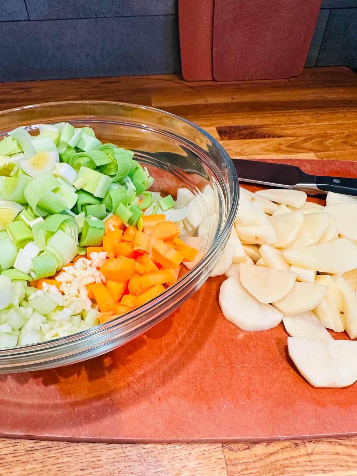Glass bowl containing chopped leek, carrot, celery, and garlic next to chopped potatoes on a cutting board.
