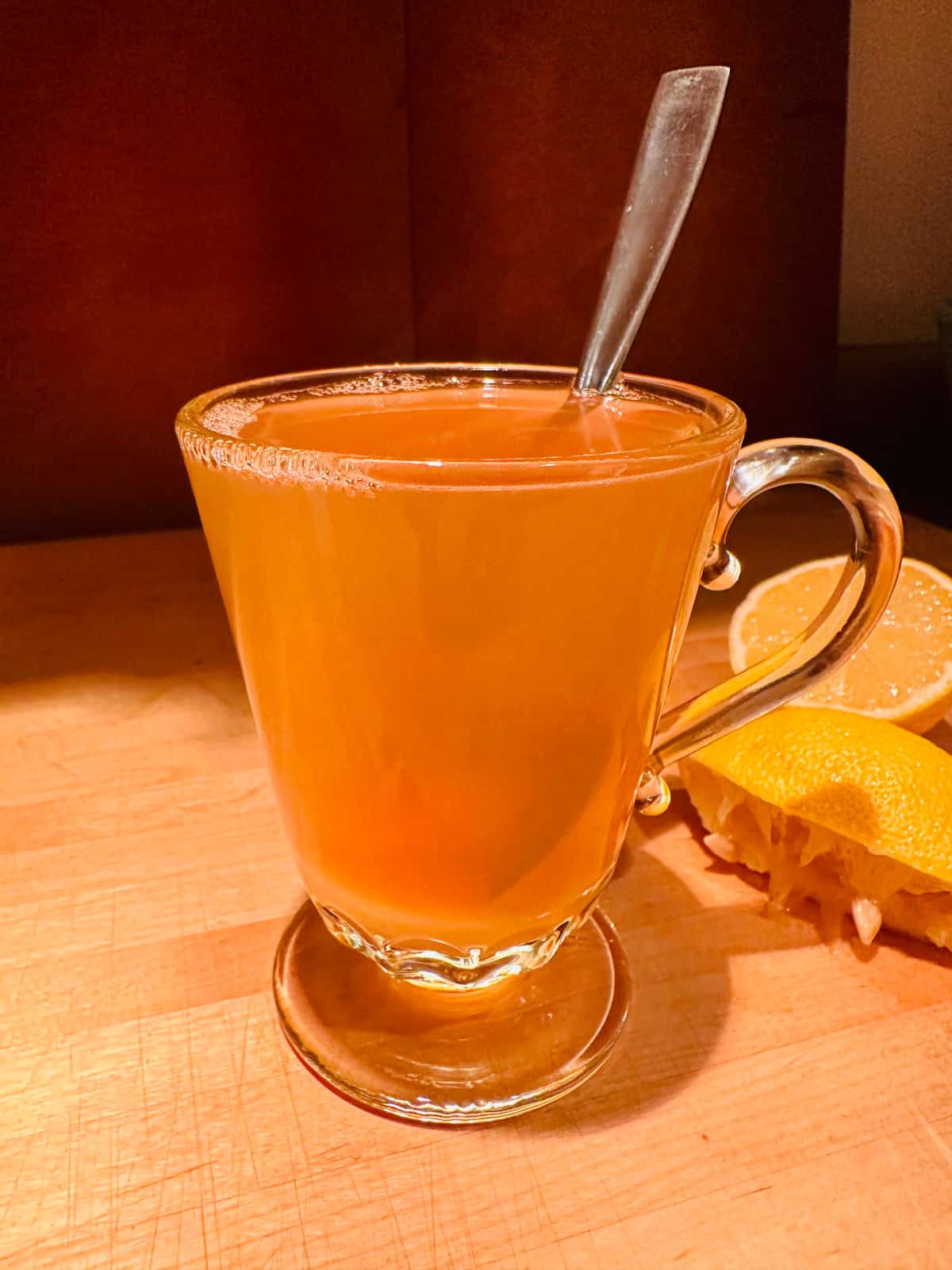 Glass mug containing honey, lemon juice, whiskey, hot water, and a spoon.