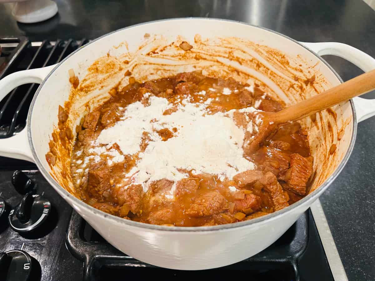 Browned beef and flour in a white pot with a wooden spoon.