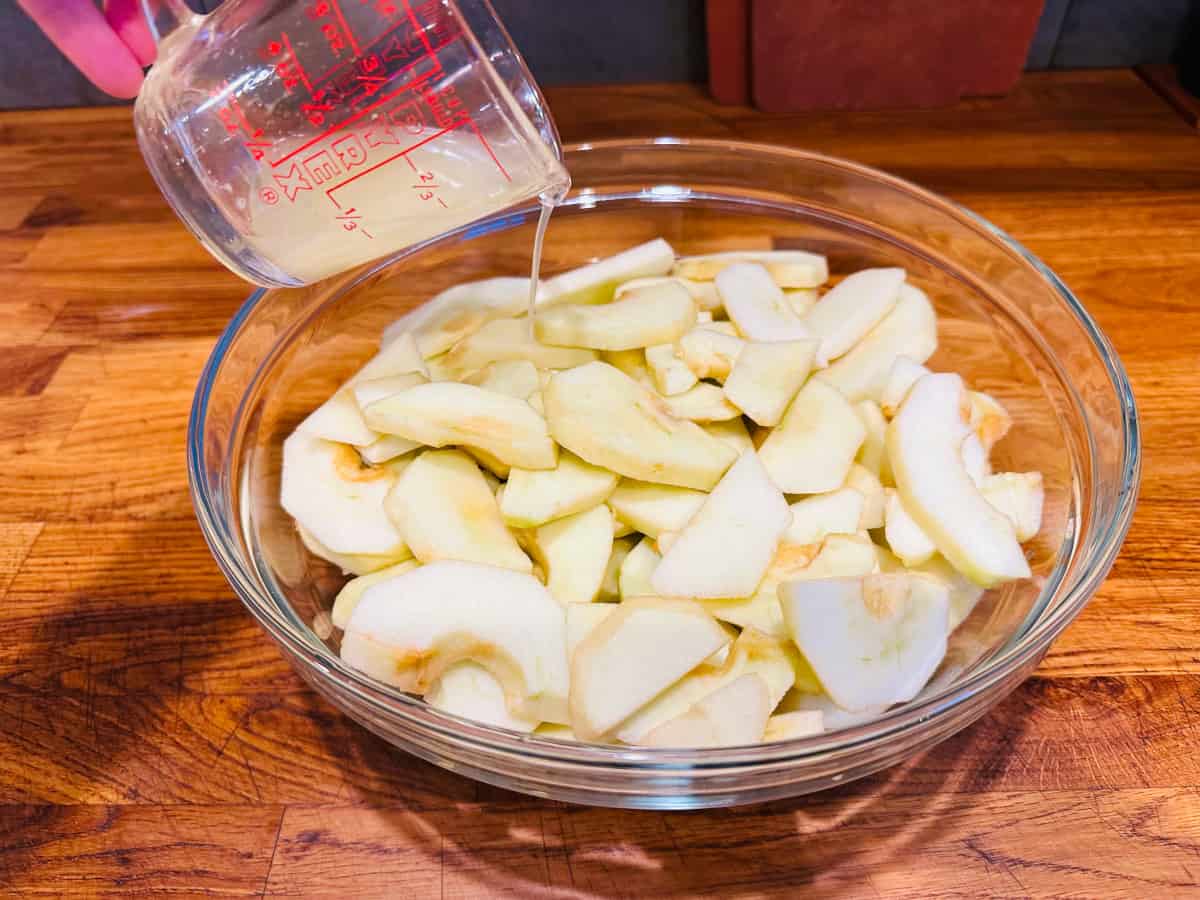 Lemon juice pouring from a pyrex measuring cup into a glass bowl full of sliced apples.